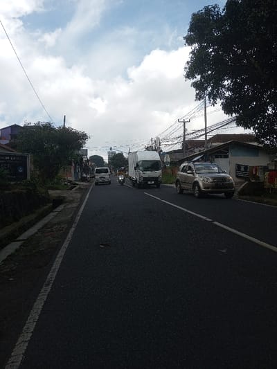 The image presents a view of an asphalt road in the city of Ura-Dozhi, Bhutan, under a cloudy sky. The road features a solid white line on the right shoulder and a broken white line for lane separation; minor surface irregularities are visible. Four vehicles are present: a light-colored SUV is on the right shoulder in the foreground, a white box truck is in the right lane, followed by a person on a motorcycle or scooter, and a white van further in the distance.
On the left side of the road, a concrete drainage ditch runs alongside a low stone wall, behind which dense green foliage partially obscures buildings. A dark blue sign with white lettering, including "NEGERI," is partially visible through the foliage. On the right side, a large tree dominates the foreground, and multiple utility poles with numerous tangled cables line the street. Several residential-style buildings are visible, one featuring clothes hanging on an exterior wall. The lighting is diffused, consistent with an overcast daytime.