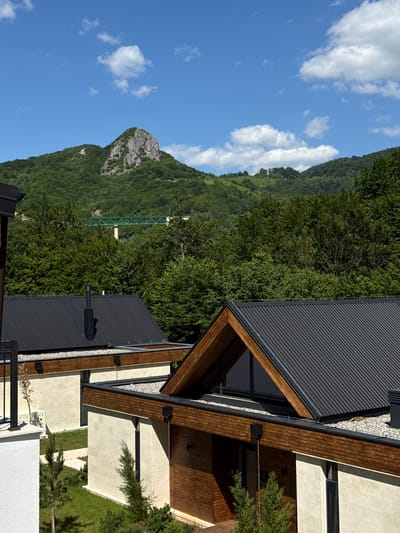 The image captures a scenic view from Bakovići, Montenegro, showcasing modern architecture nestled against a lush, mountainous backdrop. In the foreground, two contemporary houses with dark, corrugated metal roofs and light-colored stone facades are visible. One house features a prominent triangular gabled roof with exposed wooden beams. The other house, partially obscured, also has a dark metal roof and a chimney. The landscaping includes manicured green grass and small evergreen trees.
Beyond the houses, a dense forest of green trees blankets the rolling hills. In the distance, a striking rock formation juts out from the mountainside, a prominent natural landmark. A long, green truss bridge spans across a valley, partially visible through the trees, indicating infrastructure within the mountainous terrain. The sky above is a vibrant blue, dotted with fluffy white clouds, suggesting pleasant weather conditions. The overall scene conveys a sense of peaceful coexistence between modern living and the natural beauty of the Montenegrin landscape.