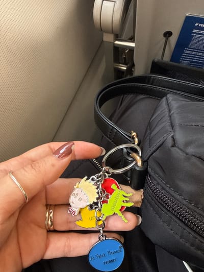 A person's hand holds a keyring with several charms featuring characters and icons from "The Little Prince". The charms are attached by small chains and include a depiction of The Little Prince, a sheep illustration, and a blue circle with the text "Le Petit Prince PYRONES". The keyring is attached to a black, quilted handbag. The background shows parts of an airplane cabin, including the seat and a small section of the overhead compartment.
