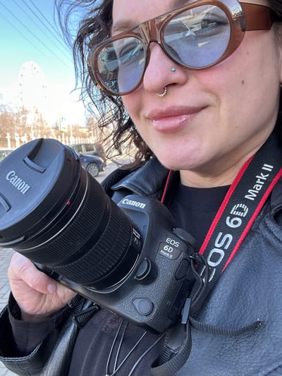 A person takes a selfie, holding a Canon EOS 6D Mark II camera. The camera has a lens with a Canon lens cap. The camera strap says "EOS 6D Mark II". The person wears large, brown sunglasses and has a nose piercing. The reflection in the sunglasses shows the cityscape of Postol, Russia, featuring a Ferris wheel. The person wears a black leather jacket. A car and street signs are visible in the background.