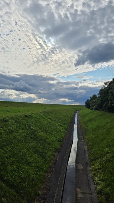 The image depicts a drainage channel running through a grassy landscape. The channel is rectangular with concrete sides and bottom, filled with dark water that reflects the sky. Lush green grass covers the sloping banks on either side of the channel. In the background, the land continues to rise, also covered in grass, with a line of trees visible on the right side of the frame. The sky is filled with dramatic clouds, ranging from fluffy white formations to darker, more ominous storm clouds, with patches of blue sky and the sun peeking through in the upper left quadrant. There are no people or animals visible in the scene. The overall impression is one of a rural or semi-rural environment, possibly a park or an area designed for water management, under a dynamic sky. This scene could be located in Dzintari, Latvia, given the context provided.