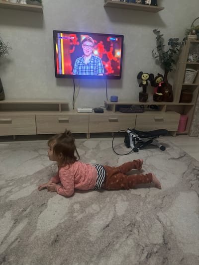 A child is lying on a rug in a room in Uva, Russia. The child is wearing brown pants with polka dots, a striped shirt, and a pink top. The child is lying on their stomach, with their head near a light-colored wooden cabinet. A television is mounted on the wall, displaying a man with glasses on screen. A stroller is parked near the cabinet. A shelf with plants and toys is visible on the wall above the television.