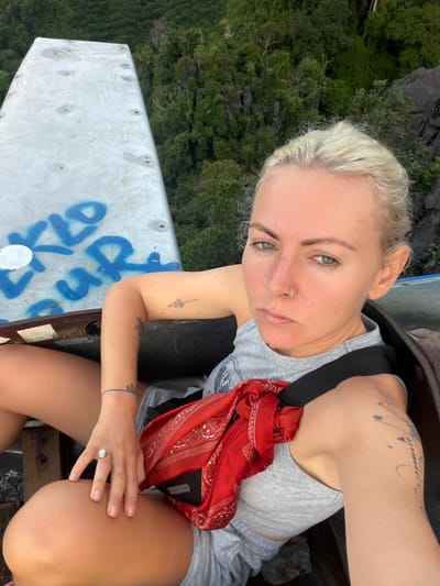 A woman with blonde hair and blue eyes is taking a selfie. She is wearing a gray tank top and gray shorts, with a red bandana tied around her chest. She has several tattoos on her arms and a ring on her finger. She is sitting on a rusty metal structure, possibly part of an old vehicle or aircraft, with graffiti on it. The background shows a lush green forest and rocky cliffs, suggesting a natural, outdoor location. The environment appears to be located on a hilltop overlooking a jungle, likely in the vicinity of Naka, Laos, given the terrain and vegetation.