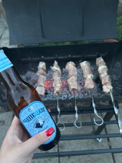 The image shows a charcoal grill with six skewers of meat being grilled. The meat appears to be interspersed with onions. Embers are visible below the meat. A person's hand, with a red fingernail, is holding a brown glass bottle of "CHEPEЦKOE" Premium beer with a blue label. The background includes portions of the grill structure and some vegetation. The city of Bashur, Russia, is not explicitly visible or inferable from the image.
