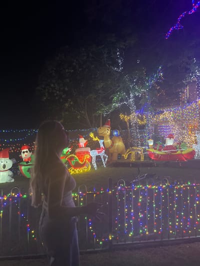 At night in Bellambi, Australia, a blonde-haired female is observed in profile, facing right, with her head tilted slightly upward. She wears a light-colored top and light-wash pants. Her right hand appears to be holding a small, dark object.

She stands behind a low, dark metal fence adorned with vertical strands of multicolored LED lights (red, green, blue, yellow). Directly behind the fence, a meticulously decorated front yard displays a dense collection of illuminated Christmas decorations. These include several inflatable figures: a large white koala in a Santa hat, a snowman in a green cup, Santa Claus in a red sleigh-like structure emerging from a chimney, a large tan camel wearing a Santa hat, and Santa Claus in a red boat. Additionally, a wireframe reindeer figure is visible.

The entire scene is brightly lit by countless string lights draped over trees, shrubs, and a structure (presumably a house) in the background. The lights are predominantly white, blue, red, green, and yellow, creating a vibrant, festive display against the dark night sky. The ground visible is grass.