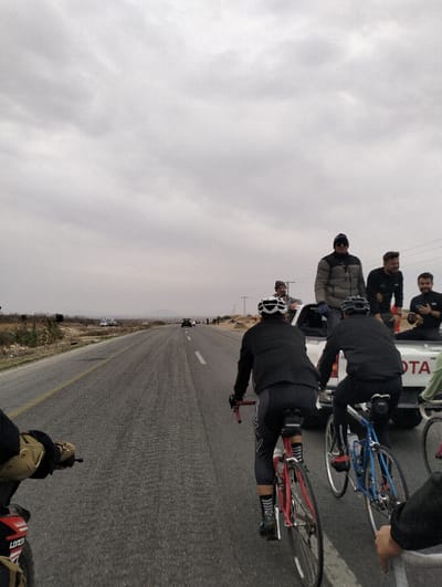 The photo depicts cyclists riding on a road with a support vehicle nearby. Several men stand in the back of a white Toyota pickup truck, some observing the cyclists. The sky is overcast. Other vehicles are visible on the road ahead. The cyclists are wearing helmets and riding gear, and the camera appears to be positioned from behind another cyclist or motorcycle.