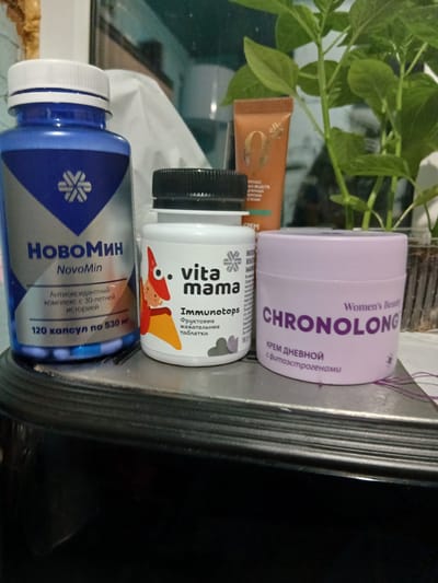 The image shows a collection of personal care products arranged on a surface, possibly a shelf or counter. From left to right, there is a blue bottle labeled "HOBOMИH NovoMin," a white bottle with a black cap labeled "vita mama Immunotops," and a lavender container labeled "CHRONOLONG." There is also an orange tube of what appears to be cream, as well as a green plant in the background. The presence of Cyrillic text on all the products suggests the location is within a Russian-speaking area. Based on the text on one of the products, the city is likely to be Ильинка, Russia.