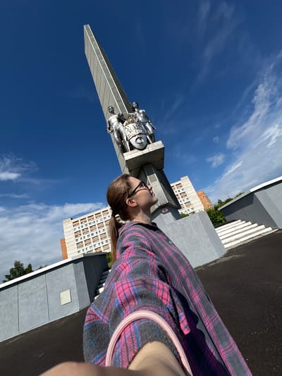 A low-angle, wide shot captures a woman in a plaid shirt looking up at a monument in Kirovo-Chepetsk, Russia. The monument, a tall, gray, obelisk-like structure, is topped with two metallic figures and a circular emblem. The woman, with her hair in a ponytail and wearing glasses, is positioned in the foreground, her upper body and face visible as she gazes upwards. Behind the monument, residential buildings and trees form the urban backdrop. The sky is a vibrant blue with scattered white clouds. The ground is paved and appears to be an outdoor public space.