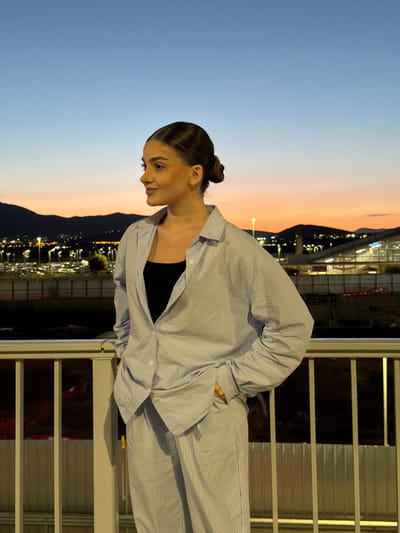 A woman stands on a balcony overlooking a city, presumably in Spata-Loutsa Municipal Unit, Greece, as indicated by the landscape and urban lighting in the distance. The time of day appears to be dusk or early evening, with a gradient of blue and orange in the sky. The woman has her hair pulled back in a bun and wears a black top under a light blue button-down shirt and striped pants. She leans slightly forward, looking down, possibly at the city below. The balcony has a metal railing with horizontal bars.