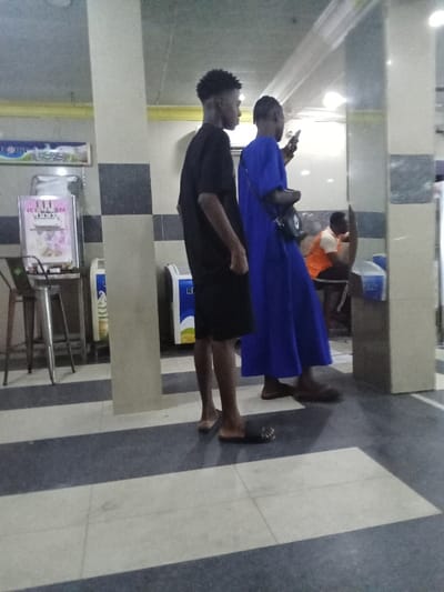 Two men stand in a tiled, indoor space. The man on the left, wearing black shorts and a black t-shirt, stands with his arms at his side looking forward. The man on the right wears a long blue robe. He is looking at his phone with his right hand while holding a black bag in his left hand. The space is sparsely furnished with a few stools and a refrigerator. There is a stainless steel ice cream machine on a small table in the background. The scene appears to be set in an ice cream shop. The tiled floor is black and grey. The ceiling is white and has recessed lights. The location appears to be in an urban setting. The coordinates, 5.503 latitude, 5.799 longitude, correspond to a location in Ovwian, Nigeria.
