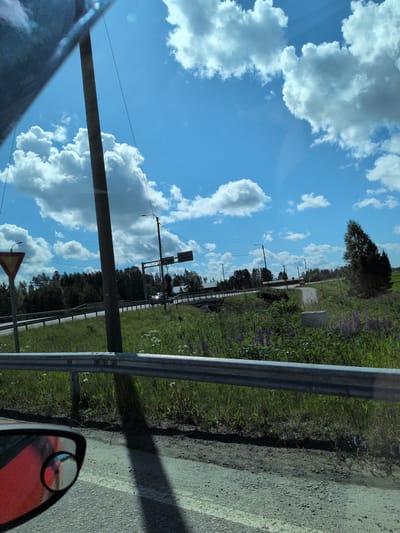 The image captures a detailed view of a rural road scene under a bright blue sky with scattered white cumulus clouds, situated in Hartola, Finland. The perspective is from inside a vehicle, with the upper left corner showing a darkened reflection of the car's interior, and the bottom left revealing part of a red car's body and its side mirror.
A weathered, grey metal guardrail runs horizontally across the lower segment of the frame. Below it, a stretch of asphalt road is visible, marked with a faded yellow line near the left and transitioning into a dirt-gravel shoulder on the right. A prominent, elongated shadow, cast by a vertical object, extends diagonally across the road surface.
The midground is dominated by a vibrant green embankment, lush with tall grass, interspersed with clusters of purple lupine flowers and smaller white wildflowers. A tall, dark wooden utility pole stands vertically, slightly left of the image center, with overhead electrical wires visible against the sky. To the immediate left of this pole, a red and yellow triangular "Give Way" road sign is partially visible.
Further into the midground, a paved road or highway is depicted, curving gently to the left and ascending onto an overpass or bridge. Multiple dark-poled streetlights line the road and bridge. An overhead gantry road sign structure spans the highway to the right of the central utility pole. A light-colored vehicle, possibly white, is observed traveling on the road beneath this overhead structure, with its headlights activated. Beyond the road infrastructure, a dense line of coniferous trees forms the background, and in the distance to the right, a few buildings with red roofs can be discerned. To the far right, a large, dark green, broadly shaped tree stands out against the sky and distant fields. The scene is illuminated by strong sunlight, creating clear shadows, indicative of a clear, sunny day.