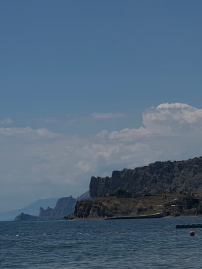 The image shows a coastal landscape in Sudak, Ukraine. The foreground is dominated by the dark blue, choppy water of the sea, with a line of orange buoys marking a swimming area. In the mid-ground, a rocky, arid coastline rises steeply. Several structures, appearing to be abandoned or in disrepair, are visible on the slope of the cliff. Further back, a series of rugged, mountainous formations form a dramatic backdrop against a clear blue sky with scattered white clouds. The overall scene is one of a remote, natural coastline with signs of human habitation.