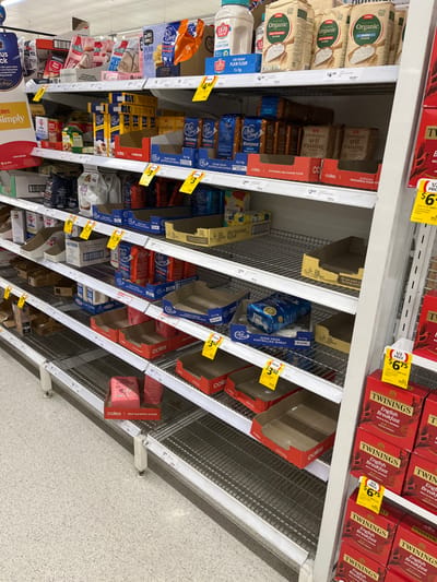 A grocery store shelf in Middle Park, Australia (latitude: -27.556, longitude: 152.923), is sparsely stocked with various grocery items. The shelves are mostly empty, with only a few boxes of self-raising flour, packets of Twinings English Breakfast tea, and a few other items remaining. The shelf is made of metal and has red and white price tags attached to it.  The shelf is tilted at an angle, suggesting that it may be in need of repair. The floor is a speckled grey tile. There is no one present.