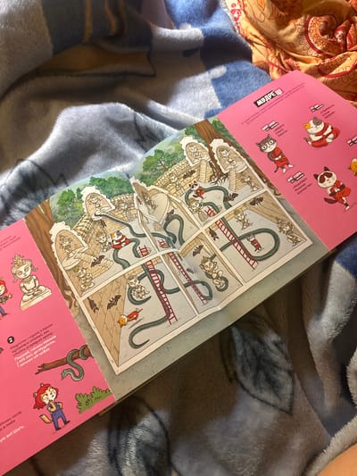 The image shows an open book with an illustration on the left and text with smaller illustrations on the right. The illustration on the left depicts a scene within a temple-like structure. Several characters, some resembling cats and others human-like figures in meditative poses, are present. Snakes and bats are also visible in the illustration. The right side of the book has text in Ukrainian, accompanied by smaller drawings of cats in various outfits. The book is placed on a blue and orange patterned blanket. The overall scene is an indoor shot with soft lighting. There is no indication of the city of Chornomors'ke, Ukraine, in the image.