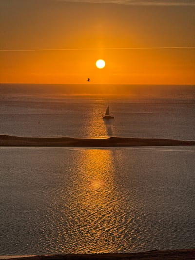 The image captures a serene seascape during sunset. The sun, a bright orange orb, is positioned on the horizon, casting a warm glow across the sky and the water. A thin, white contrail stretches horizontally across the upper portion of the sky, and two dark silhouettes of birds are visible in flight. In the middle distance, a small sailboat with a dark sail navigates the calm sea, with its reflection shimmering on the water's surface. A thin, elongated sandbar or landmass separates the middle section of the sea from the foreground. The water in the foreground reflects the vibrant orange of the sunset, creating a dazzling display of light and color. A solitary, dark figure of a person stands at the very bottom edge of the frame, on what appears to be a sandy incline. The scene evokes a sense of tranquility and natural beauty. This image was captured at the Dune du Pilat, also known as the Dune of Pyla, located in La Teste-de-Buch, France, a popular tourist destination renowned for its immense sand dune and panoramic views of the Atlantic Ocean and the Arcachon Bay.