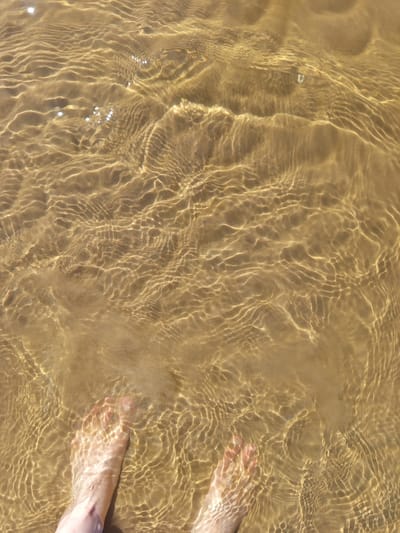 The image depicts a high-angle view of a person's feet submerged in shallow, clear water over a sandy bottom. The location is Podborov'e, Russia. The water surface exhibits significant ripples, causing intricate, undulating patterns of refracted sunlight to be cast upon the light brown sand below. These light patterns vary in intensity and shape across the visible area. The person's bare feet, specifically the anterior portions, are visible at the bottom of the frame, partially obscured by the water and its refractive effects. The skin tone of the feet appears light. No other human figures, objects, or significant environmental details are present beyond the water, sand, and feet. The overall illumination suggests bright, direct sunlight.