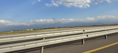 The image shows a view from a moving vehicle on a highway in the city of Zharmukhabmet, Kazakhstan. In the foreground, a silver metal guardrail runs parallel to the asphalt road, with a yellow line marking the edge of the lane. Beyond the guardrail, a wide, flat plain stretches towards a distant mountain range. The mountains are capped with snow and shrouded in a light haze. A few buildings and structures are visible on the plain, suggesting some development in the area. The sky is a clear blue with scattered white clouds. The overall scene depicts a vast, open landscape with a highway cutting through it.