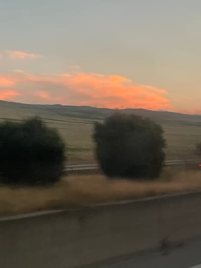 A blurred, horizontal, eye-level shot depicts a landscape at dusk. The sky is a gradient of soft blue and peach, with wispy, orange-tinged clouds. Below the sky are rolling hills covered in green fields. In the foreground, two dark, bushy trees obscure a portion of the landscape. A highway is visible between the trees, with a few distant lights suggesting vehicles. The lower part of the image is dominated by a dark, blurred foreground that appears to be a road surface. The overall impression is one of a serene, natural setting at the end of the day. The image does not contain any recognizable landmarks or features that would allow for specific location identification within the city of Dzveli Kanda, Georgia.