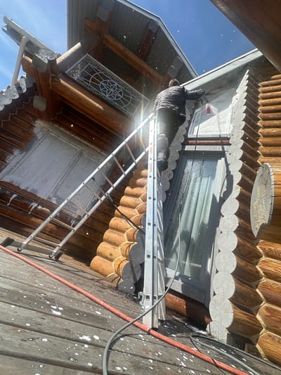 A person is on a ladder, working on the exterior of a log cabin-style building in Areyskoye, Russia. The person is wearing dark clothing and appears to be cleaning or painting the building. The ladder is positioned against the wall, and there is an orange hose and a gray cable in the frame. The building has a window with a gray frame. The sky is blue. The exterior of the cabin shows signs of paint splatter.