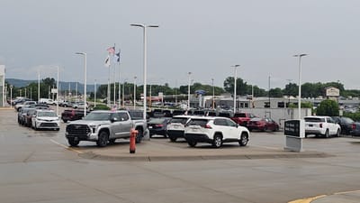 The image shows an outdoor car dealership in Winona, United States of America. Numerous cars, predominantly SUVs and trucks, are parked in rows on a wet asphalt lot. In the foreground, a silver Toyota Tundra pickup truck is parked, with a red fire hydrant next to it. To the right, a white Toyota RAV4 SUV and a white Toyota Highlander SUV are visible. Further back, other vehicles are parked, including sedans and more SUVs. In the background, a sign for "Winona" with a logo is visible on a building, along with a Ford dealership sign. The sky is overcast, and there are trees and distant hills in the background. Flags, including the American flag, are flown on tall poles.