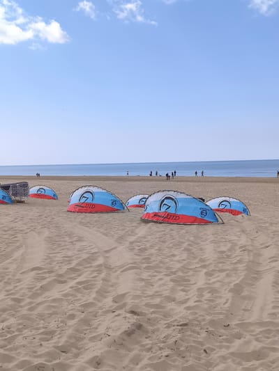 This image depicts a beach scene in Bergen, Netherlands. The foreground is dominated by a sandy beach with distinct tire tracks. Scattered across the beach are several windbreaks, each bearing a logo that reads "7 LUID." A stack of chairs sits to the left. In the midground, a group of people are visible near the waterline. In the background, the sea meets a clear blue sky with a few clouds. The weather appears sunny and bright.