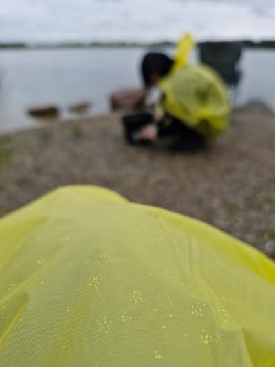 A close-up, low-angle shot focuses on the textured surface of a bright yellow raincoat, possibly with water droplets or debris. In the blurred background, a person wearing a yellow poncho is crouched near a body of water, appearing to be engaged in an activity involving a dark bucket. The environment suggests an outdoor setting near a lake or river, with a pebbly shoreline and a cloudy sky. The image is taken in Gargždai, Lithuania.