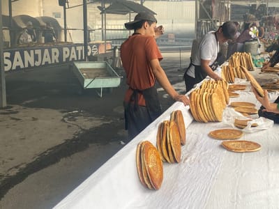 This image depicts an outdoor market scene in the city of Pungon, Uzbekistan, likely a bakery or a stall selling freshly baked bread. The foreground is dominated by a white cloth-covered table laden with numerous round, golden-brown flatbreads, some stacked and others individually placed. These flatbreads exhibit decorative patterns pressed into their surfaces.
In the mid-ground, two men are visible. One, wearing a dark baseball cap and an orange t-shirt with a black apron, stands facing away from the camera, appearing to be working or overseeing the bread. The other man, wearing a baseball cap and a white t-shirt with a black apron, is bent over the table, interacting with the bread. Behind them, a large blue sign with white lettering that reads "SANJAR PATI" is mounted on a structure. Further in the background, ovens or kilns are visible, suggesting the production site for the bread. The ground is a dark, textured surface, possibly concrete, with some dark stains or moisture. The overall atmosphere appears to be that of a busy marketplace with food being prepared and sold.