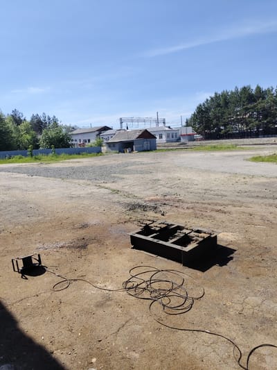 The image depicts an outdoor industrial or service area in Shimanovsk, Russia, under a clear blue sky. In the foreground, a dark metal rectangular frame with internal structures rests on the ground. To the left, a smaller, dark metal object, possibly a support or stand, is connected by a thick black cable that coils loosely on the dirt and concrete surface. The ground is a mixture of dirt and cracked concrete, with scattered debris. In the middle ground, a blue metal fence runs horizontally, separating the foreground area from a gravel and dirt expanse. Beyond the fence, several low-rise buildings with grey roofs are visible, one with a small brown-roofed structure attached. Trees line the background, with a clear blue sky above. There are no people or animals in the image. The scene appears to be a storage or work area for equipment, possibly related to railway or industrial maintenance given the presence of what appear to be railway tracks and overhead power lines in the distance.