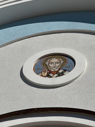 A mosaic icon of Saint Nicholas is embedded in the exterior wall of a building, which is located in the city of St. Ozerki, Russia. The mosaic is circular and framed by a white, oval-shaped protrusion from the building's facade. The saint is depicted with a halo and is wearing a priestly vestment. The background of the mosaic is blue, and the saint's clothing is detailed with geometric patterns in various colors. The building's exterior is textured and painted in light colors, with a curved, light blue band above the icon. The sunlight casts a shadow to the right of the mosaic, indicating the sun is to the left.