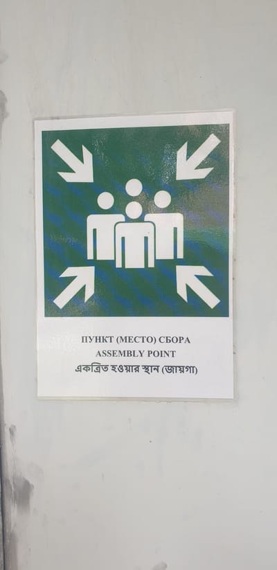 The image shows a safety sign for an assembly point, a designated area where people should gather in case of an emergency. The sign is white with a green background and features four white arrows pointing inwards towards a group of three stylized human figures. The text below the symbol includes "ПУНКТ (МЕСТО) СБОРА", "ASSEMBLY POINT", and text in Bengali script, "একত্রিত হওয়ার স্থান (ভয়গা)". The sign is mounted on a light-colored wall with some marks and imperfections. This sign is located in Rooppur, Bangladesh, likely within the Rooppur Nuclear Power Plant complex.