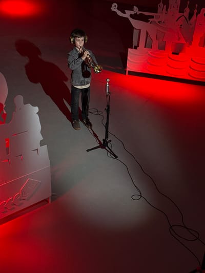 A young male musician is captured in a high-angle, tilted photograph, performing indoors in a scene dominated by dramatic red lighting. Located in Kurchatov, Russia, the musician, wearing a dark denim jacket, dark pants, and brown shoes, is equipped with over-ear headphones. He is actively playing a gleaming gold-colored trumpet, positioned near a silver condenser microphone on a black stand. Black cables snake across the floor or surface.

The intense red light emanates from the left and bottom right, casting a long, distinct shadow of the musician behind him. The backdrop or surrounding environment features large, stylized white or light-colored cutouts. On the left side, multiple layers of geometric cutouts clearly spell out "ABBA," repeated and stacked for a three-dimensional effect, alongside a cutout of a stylized human figure, possibly an athlete or dancer. Another cutout with geometric patterns is visible in the bottom right corner of the frame. The musician appears focused on his performance. The setup suggests a concert, rehearsal, or studio recording session.