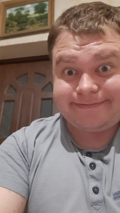 The image is a selfie of a man taken indoors. The man has light skin and brown hair. He is wearing a light gray t-shirt with buttons. He has a surprised expression with wide eyes and his mouth is slightly open. In the background, to the left, is a framed painting of a landscape. Behind the man, to the left, is a wooden door with decorative panels. The setting appears to be a domestic interior. There is no information about the specific location in Vatra, Moldova, in the image itself.