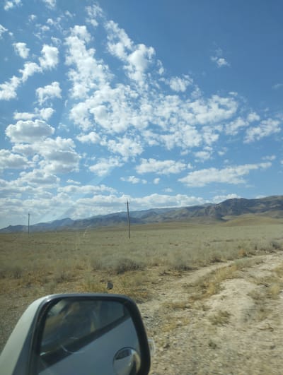 The image shows a view of a vast, arid landscape with low, rolling hills in the distance under a bright blue sky filled with fluffy white clouds. In the foreground, a dirt road or track curves through dry, sparse vegetation. A side-view mirror of a vehicle is visible in the lower left corner, reflecting part of the vehicle's interior. Two utility poles with wires are sparsely placed in the landscape, indicating some form of infrastructure. The overall scene is one of a remote, undeveloped area. The location is stated to be in the city of Temirkavuk, Uzbekistan.