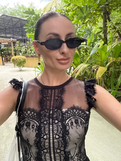 An outdoor, eye-level, medium selfie shows a woman with dark hair pulled back, wearing large, oval-shaped black sunglasses, a black lace top with ruffled sleeves, and dangling earrings. She is positioned in the center of the frame, looking at the camera. Her lips are painted with a glossy nude lipstick. A silver necklace is visible around her neck. A white and black strap of a bag is draped over her left shoulder. The background features lush green tropical foliage and trees, with a glimpse of a wooden structure and some tables and chairs to the left. The ground is a light green paved surface. The scene appears to be in a garden or resort area.