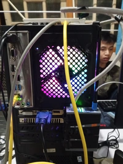 The image displays the rear of an active desktop computer setup within an indoor environment in Al Haouafate, Morocco. A black computer tower is centrally positioned, with its internal RGB-illuminated fan visible through a circular mesh grille, cycling through pink, purple, green, and blue colors. The rear I/O panel features multiple connected cables: a black cable in a USB port, a yellow Ethernet cable with active green and amber indicator lights, and a blue VGA connector plugged into its corresponding port. A gray cable and additional black cables are also visible, traversing the scene. A dark gray, stacked multi-port adapter with active green lights is attached to the right side of the computer tower, into which a blue connector is plugged. In the blurry background, to the right of the computer tower, a young male with short dark hair, wearing a patterned garment, is partially visible, observing something off-frame to the lower right. A dark-colored keyboard rests on a surface to the right of the male. Behind the computer, a blurry window or framed opening with horizontal bars is discernible. The setup includes multiple cables on the desk surface.