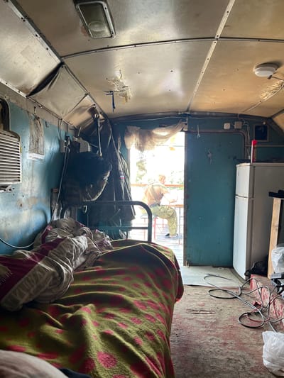 The image depicts the interior of a small, cluttered room, possibly a sleeping compartment or a makeshift living space. A bed, covered with a green blanket featuring pink polka dots and a red and white striped blanket, is prominent in the foreground. The walls and ceiling appear to be made of metal panels, some showing signs of wear and tear. Various items are scattered around, including clothing hanging on hooks, a refrigerator, electrical cords on the floor, and what looks like a small air conditioning unit on the left wall.
In the background, a person is seated at a table near an open doorway, looking outwards. The person is wearing camouflage pants and a light-colored t-shirt. Sunlight streams in through the doorway, illuminating the space outside, which appears to be an outdoor area with some greenery. The overall impression is one of a utilitarian and somewhat spartan living environment. The image does not contain any specific visual elements that allow for the localization within the city of Koshara, Russia.