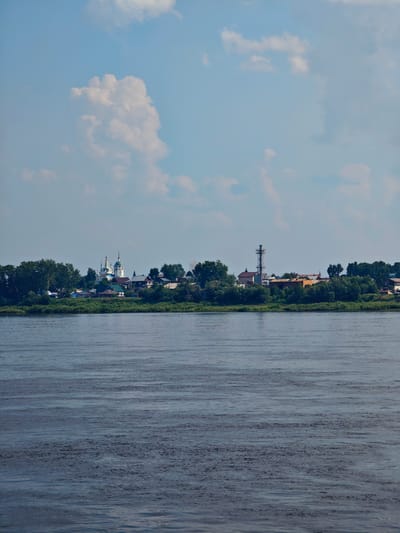 The image depicts a wide river in the foreground, with gentle ripples disturbing its surface. On the far bank, a town is visible, nestled behind a lush green shoreline and dense trees. The town features a prominent white church with blue domes and spires, suggesting a religious or historical significance. Adjacent to the church are several buildings, including residential homes and what appears to be industrial structures with a tall, metal tower. The sky above is a pale blue, dotted with fluffy white clouds, indicative of a partly cloudy day. The overall impression is that of a serene, rural or semi-urban landscape. The town shown is likely located in Yeniseysk, Russia, given the characteristic architecture and landscape often associated with the region. There are no people or animals visible in the frame.