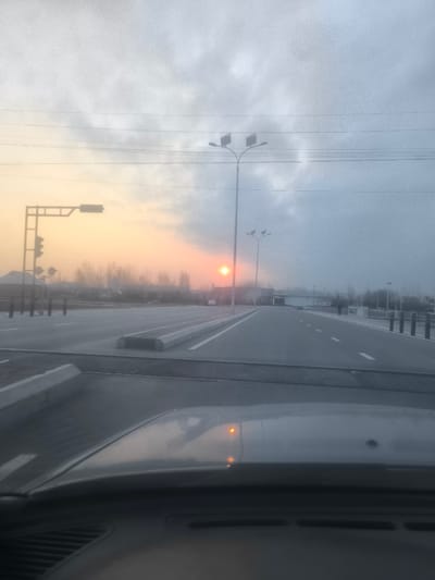 The image depicts an overcast and hazy roadside scene in Pakhtakor, Uzbekistan, at either sunrise or sunset, with a distinct orange sun low on the left horizon. The perspective is from inside a vehicle, with the grey hood and two amber indicator light reflections visible in the lower foreground. The central subject is a multi-lane asphalt road, divided by concrete median barriers and marked with white lane lines.
On the left side of the road, a gantry-style traffic light structure with multiple signal heads is visible, alongside black cylindrical bollards. Further down this section, a single white passenger car is visible in the distance. Tall streetlights with twin-fixture heads are positioned along the road, one prominently on the left and another further down the road on the right.
On the right side of the road, a pedestrian is observed walking in the distance near the horizon line. Bare trees and low-lying buildings are faintly discernible on the distant horizon, obscured by the atmospheric haze. Power lines traverse the upper portion of the frame against the cloudy sky. No direct interactions between individuals or vehicles are clearly depicted. The overall environment is one of an open, possibly semi-urban or highway intersection, characterized by infrastructure and low visibility due to atmospheric conditions.