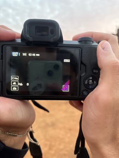 A pair of adult hands holds a black digital camera, viewed from its rear, in Nong Ian, Laos. The camera features a rear LCD screen and an electronic viewfinder. The left wrist displays a multi-strand bracelet with small, gold-colored beads. The right thumb exhibits a superficial mark near the cuticle. The camera's active LCD screen displays digital interface elements, including "A+", "59", "02:46", a near-full battery indicator, and icons related to flash, drive mode, and vehicle access (car with "OPEN"). "Exp.SIM" and a "Bluetooth OFF" indicator are also present. A distinct reflection of a dark green or gray smartphone is visible on the screen, along with a blurred white circular object beneath it. A stylized purple and red icon is displayed in the bottom right of the screen. The background, visible around and through the camera's display, is a heavily blurred outdoor scene, showing a light, possibly overcast sky at the top and a reddish-brown, unidentifiable ground surface below. A black camera strap is partially visible at the bottom.