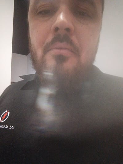 The image is a close-up, low-resolution photograph of a person's face and upper torso. The person appears to be male, with a beard and stubble. He is wearing a black shirt with a logo on the left side. The logo is a white oval with a red lightning bolt inside. Below the logo, there is white text that is partially obscured, but appears to read "20". The background is a wall that is light colored on the right and dark on the left, with a corner in the middle. The image is blurred and grainy, and there is a significant light reflection or glare across the middle of the image, obscuring details of the person's chin and neck. There is no information in the image to indicate it is located in Ümraniye, Turkey.