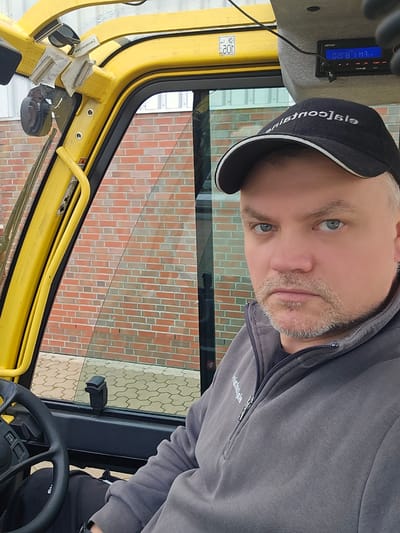 A man wearing a black baseball cap and a grey zippered sweatshirt is sitting in the driver's seat of a yellow forklift, looking at the camera. The forklift cab is partially visible, with a yellow roll bar and a window. Outside the window, a brick wall is visible. The man's face is in focus, and he has blue eyes and a grey beard. A digital display is visible above the man's head, showing the numbers "02.58 FM 3". The image appears to be taken in Haren (Ems), Germany, based on the presence of the name "Haren" on a sticker on the forklift.