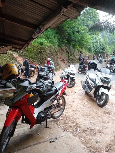 The image shows a dirt road lined with motorcycles in Cisewu, Indonesia (latitude: -7.41, longitude: 107.542). The road is situated beneath a steep hillside covered in dense foliage. There are at least six motorcycles parked along the road. One of the motorcycles, a red and white Suzuki, is positioned in the foreground, facing left, with a black bag on the back seat. There are several other motorcycles behind it, including a white Yamaha and a blue motorcycle. Two people, one wearing a dark jacket and another in a black and yellow jacket, stand near the motorcycles. The scene is partially obscured by a wooden overhang on the left, possibly part of a house or building. There are several other people walking along the road.