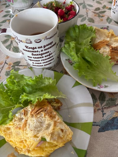 A breakfast scene is laid out on a table covered with a patterned tablecloth. In the foreground, a white plate with a green and white striped rim holds a folded omelet topped with fried crispy bits and a serving of fresh green lettuce. To the left of this plate, a white mug with brown text and drawings, containing dark liquid, is partially visible. The text on the mug appears to be in Russian and includes the word "Americano." Behind the mug, a small bowl filled with fresh strawberries is visible. Further to the right, another white plate with a floral pattern and gold trim holds slices of bread and a portion of lettuce. The setting suggests a meal, possibly breakfast, in a domestic environment. There is no visible indication of the city of Shipovo, Russia, in the image.