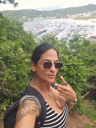 The image displays an adult female in the foreground, situated on a verdant hillside overlooking a bay in Arraial Do Cabo, Brazil. The individual, with dark hair, wears octagonal sunglasses, a black and white striped V-neck top, and a black backpack. Visible tattoos, featuring floral and leafy designs, are present on her left upper arm. She accessorizes with ornate dangling earrings, a silver-toned necklace bearing a pink crystal pendant, a silver bracelet on her left wrist, and a prominent ring with a dark green stone on her right ring finger. Her right hand is raised, forming a two-finger upright gesture. The immediate foreground shows dense green foliage and reddish-brown earth. The midground features an expansive bay, characteristic of Arraial Do Cabo's harbor, filled with dozens of moored boats of various sizes, many docked at a pier. A light-colored sandy beach is visible along the bay's edge, with several yellow inflatable vessels present on the sand. The background comprises the townscape of Arraial Do Cabo, with numerous buildings extending along the distant shoreline and climbing the surrounding hills, which are densely covered in green vegetation. The sky overhead is uniformly overcast.