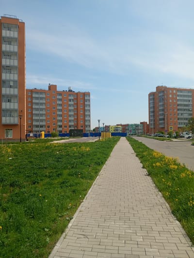The image shows a residential area in Kommunar, Russia, on a clear, sunny day. In the foreground is a light grey brick-paved walkway stretching into the distance. On either side of the walkway is a patch of green grass with yellow wildflowers. Flanking the walkway are several orange apartment buildings of similar height, with multiple rows of windows. A few black streetlights are situated between the buildings. Beyond the first set of buildings, several colorful buildings can be seen in the background. A parking lot to the right shows several parked cars. A blue construction fence surrounds the base of the buildings, and several green trash bins stand near the buildings.