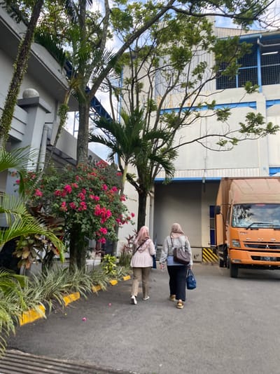 Two individuals, identified as adult females wearing hijabs, walk away from the camera on a paved asphalt path in what appears to be an industrial or commercial complex within the city of Cipeundeuy, Indonesia. The individual on the left wears a patterned light-colored hijab, a light-colored top, and light-colored pants. The individual on the right wears a beige hijab, a light blue top, a dark jacket, dark pants or a skirt, and carries a dark brown shoulder bag and a dark blue handbag. They are walking towards a large, light-grey building with blue structural accents, specifically towards a loading bay area visible on the right.
To the right of the walking individuals, an orange delivery truck with a blue stripe on its cargo box and a white license plate (partially visible as "D 8258 FF") is parked, angled slightly towards the camera. The environment features well-maintained landscaping, including a prominent bush with vibrant pink flowers (Bougainvillea) and various green plants and palm trees lining the pathway, bordered by yellow-painted curbs. Large trees with green foliage are scattered throughout, some partially obscuring the buildings. Vines grow on the grey, multi-tiered facade of a building on the far left. The sky is overcast or partly cloudy, indicating daytime.