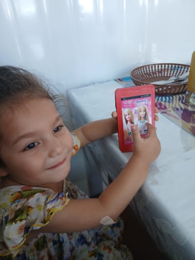A young girl with dark hair and brown eyes is holding a red smartphone in the city of Gulistan, Uzbekistan. She is wearing a light-colored dress with a dinosaur and palm tree print and a yellow trim. The girl is smiling and looking to her right. The smartphone's screen displays two Barbie dolls and the time 12:00. The background consists of a white wall and a table covered with a white tablecloth. A woven basket filled with cutlery is visible on the table.