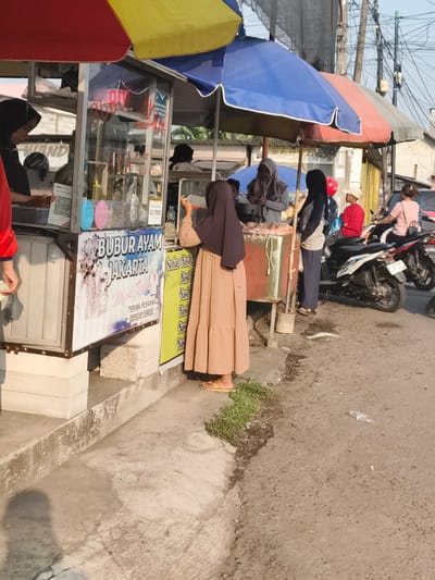 The image depicts a street scene in Barengkrajan, Indonesia, likely in the morning or late afternoon due to the soft lighting. A food stall selling "Bubur Ayam Jakarta" is the central focus. A woman wearing a beige hijab and a long, beige dress is standing at the stall, seemingly interacting with the vendor or preparing to receive food. Another woman, also in a hijab, is visible behind the counter, tending to the stall.

Several motorcycles are parked nearby, with one being ridden by a woman in a pink shirt, suggesting a bustling commercial area. A young boy in a red shirt and a light-colored cap is also present. The stall itself has a colorful awning and displays various food items and signs, including a QR code. The ground is a mix of concrete and asphalt, with some patchy grass and debris, indicating a typical urban environment. The overall atmosphere is that of a busy street food vendor operation.