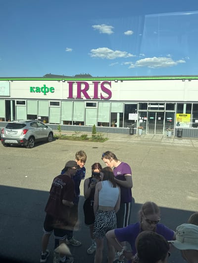 The image shows a group of people standing on a paved surface next to a building. The building has the word "IRIS" written vertically in large letters, with the word "KAFE" (cafe) below it in smaller letters. The building appears to be a business, possibly a cafe or store. A silver car is parked in front of the building. The sky is visible in the background, and there are some clouds. The image was likely taken in Казань, Russia, based on the Cyrillic lettering of the license plate of the car. The group of people seems to be looking at something or someone in front of them.