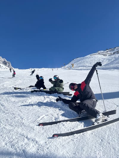 The scene depicts a brightly lit, snow-covered ski slope in Klösterle, Austria, under a clear blue sky. Three individuals are prominently positioned in the foreground and mid-ground, appearing to be in a low crouch or seated stance on their skis. The subject on the right wears a black and red ski suit, black helmet with red-tinted goggles, and black gloves; their left arm is extended upward holding a ski pole, while their right hand is open towards the snow. Attached to their feet are black skis featuring a small red and white flag emblem near the tail. To their left, a second subject in a dark ski suit, black helmet, and blue reflective goggles has their left hand raised. Further left, a third subject in an olive green ski suit, black helmet, and silver reflective goggles extends both hands with thumbs-up gestures. In the background, numerous other skiers are visible across the wide, ascending slope. A jagged, snow-dusted mountain peak is present on the far left horizon, and a series of snow fences along with several yellow structures, consistent with snow-making machines or lift components, line the right side of the upper slope.